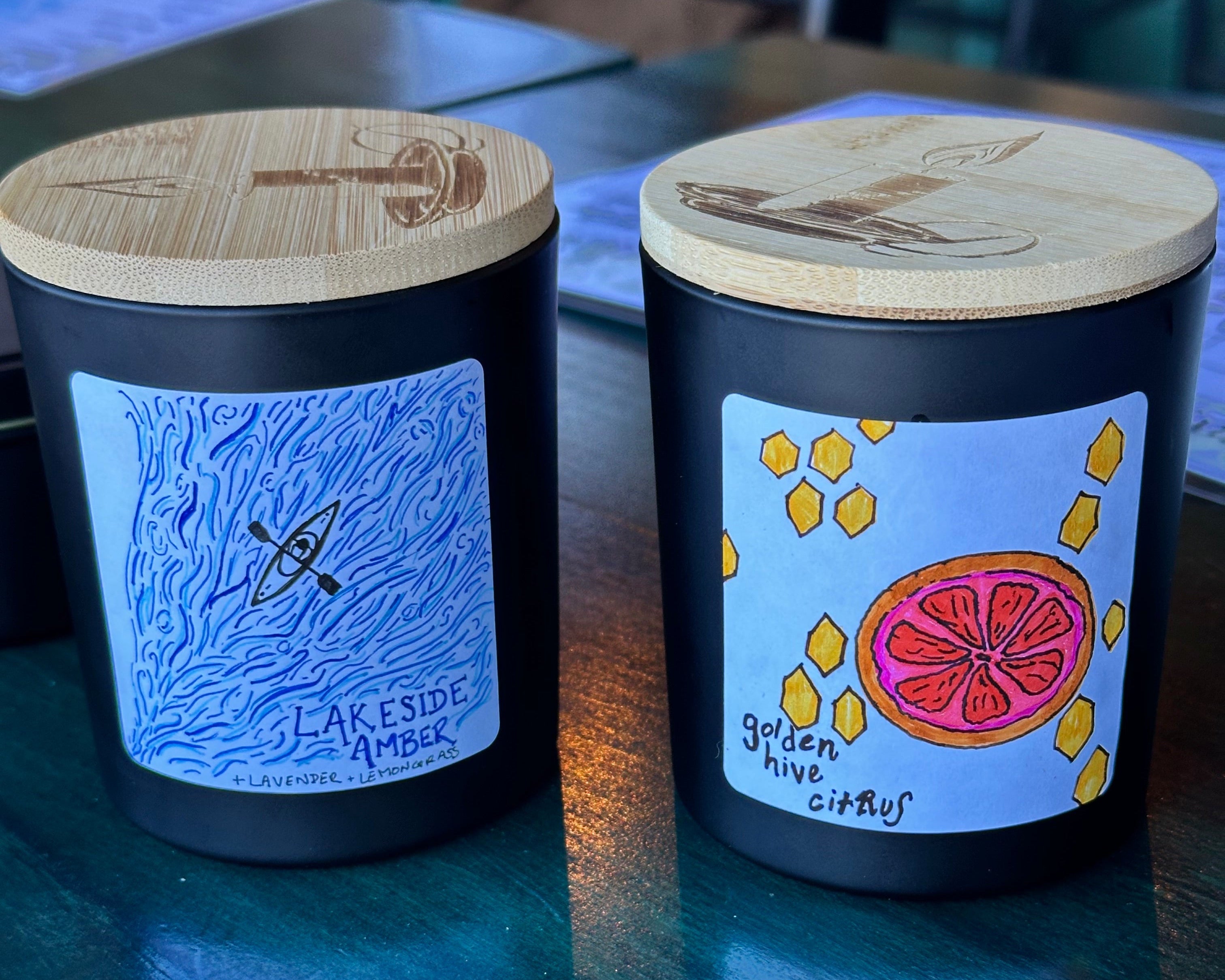 Two black canlde containers with wooden lids on a reflective surface.